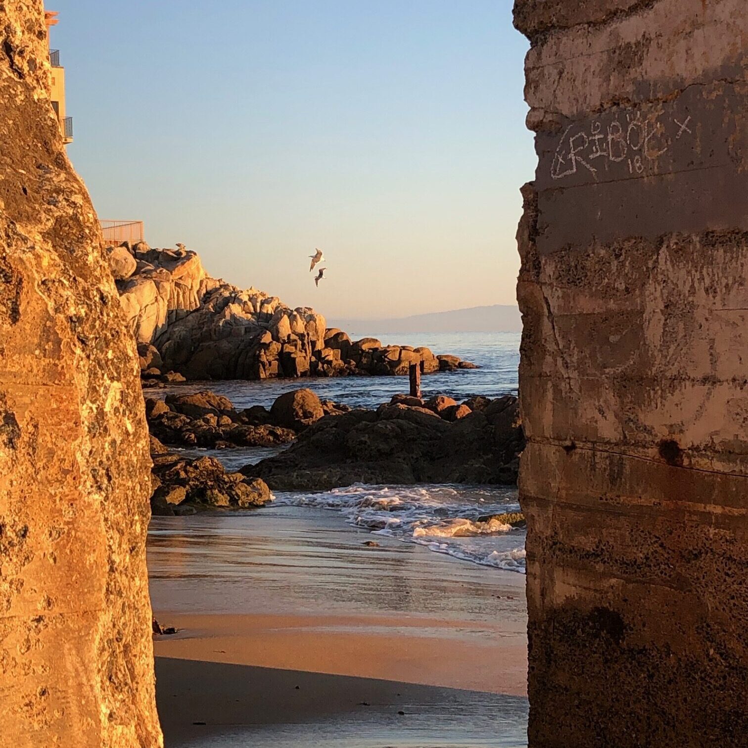 Sunset view of rocky beach through a narrow gap in stone walls.